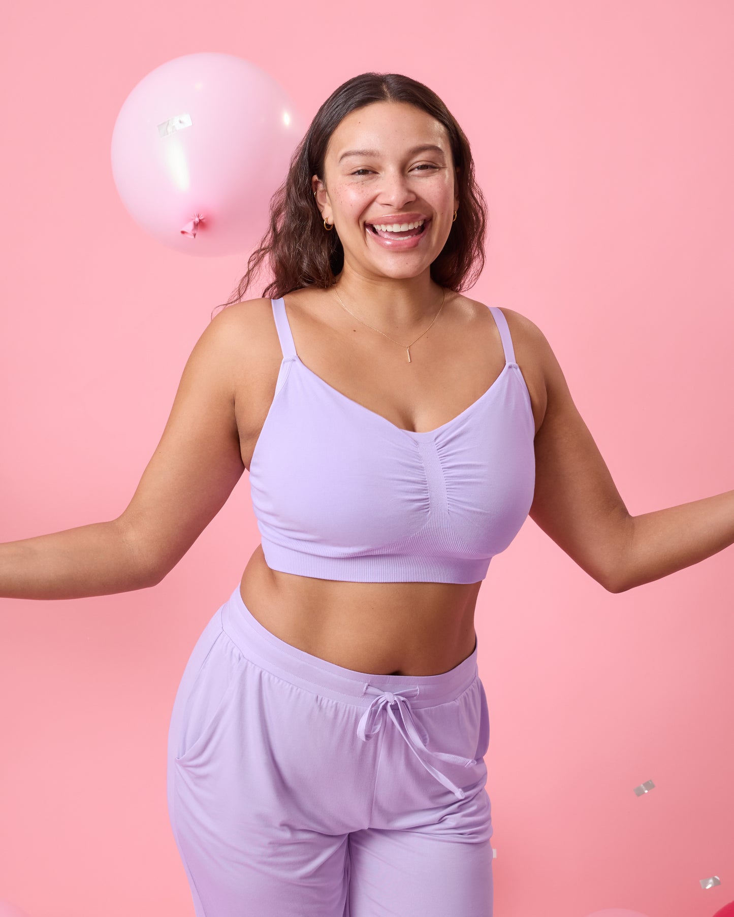 Front view of model wearing Sublime Bamboo Hands-Free Pumping Lounge & Sleep Bra in Lavender on a pink background with balloons and confetti falling around