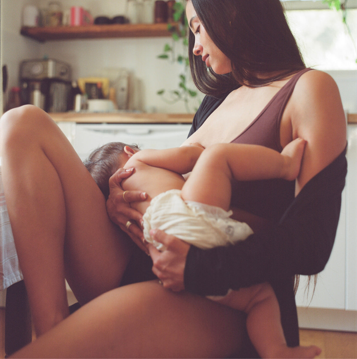 Model is breastfeeding baby and wearing Every Stage Crossover Bra in Cocoa and Every Stage Brief in Black with a kitchen background.
