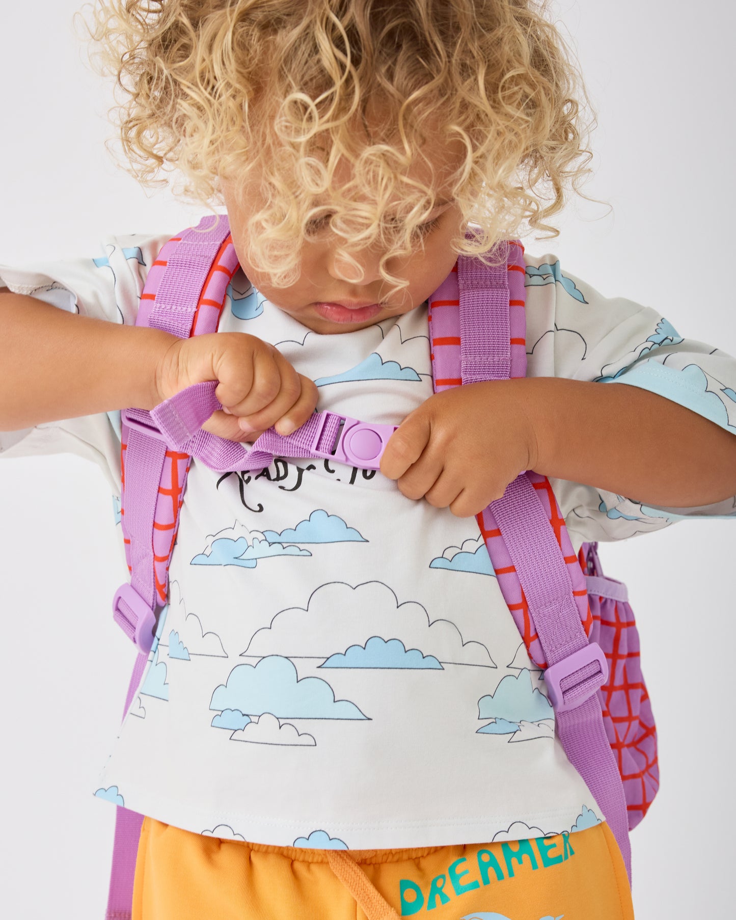 Child is clipping together shoulder straps of the Cool Kid Backpack in Orchid Grid on a beige background.