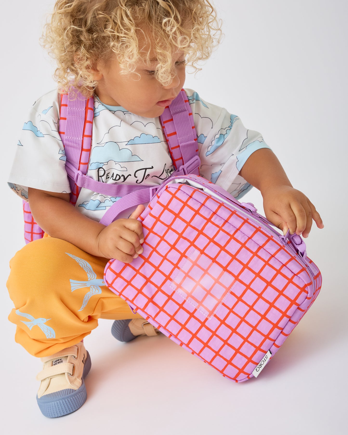 Child is kneeling down and opening the Cool Kid Lunch Box in Orchid Grid while wearing the Cool Kid Backpack in Orchid Grid on a beige background.@model_info:Waylon is 3 years old.