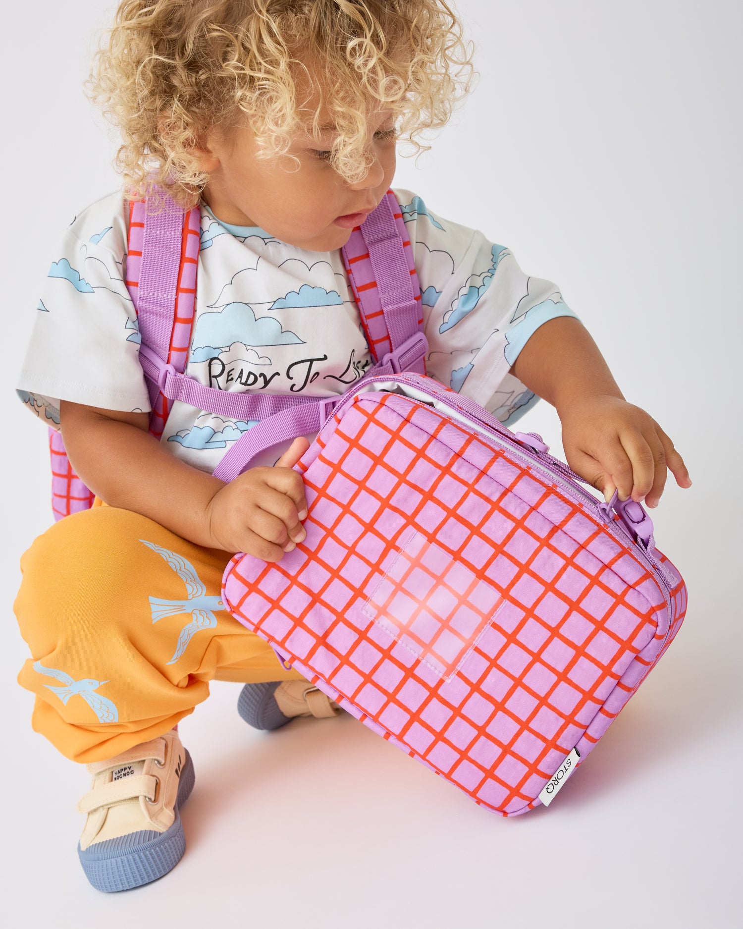 Child is kneeling down and opening the Cool Kid Lunch Box in Orchid Grid while wearing the Cool Kid Backpack in Orchid Grid on a beige background.@model_info:Waylon is 3 years old.