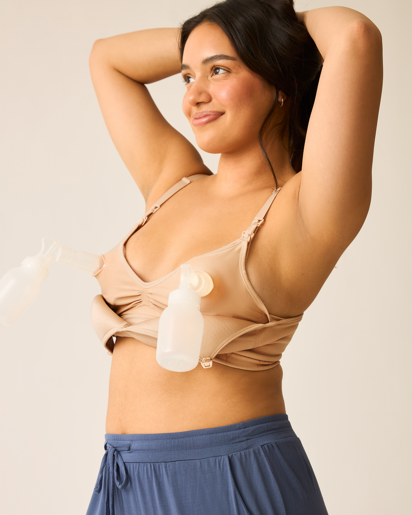Woman with hands behind head wearing the beige Signature Sublime® Contour Hands-Free Pumping & Nursing Bra with two pump flanges against a neutral background@model_info:Amber is wearing a Medium.