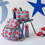 Cool Kid Lunch Box in Blue Apple on a chair next to Cool Kid Water Bottle in Blue Apple and Cool Kid Backpack in Blue Apple, two giant pencils and a pair of red sandals, on a beige background decorated with playful star and bird shapes.