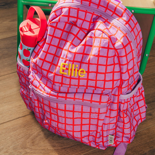 Cool Kid Backpack in Orchid Grid sitting on floor by chair, with embroidered name on front and Cool Kid Water Bottle in Blue Apple, in the side pocket. 