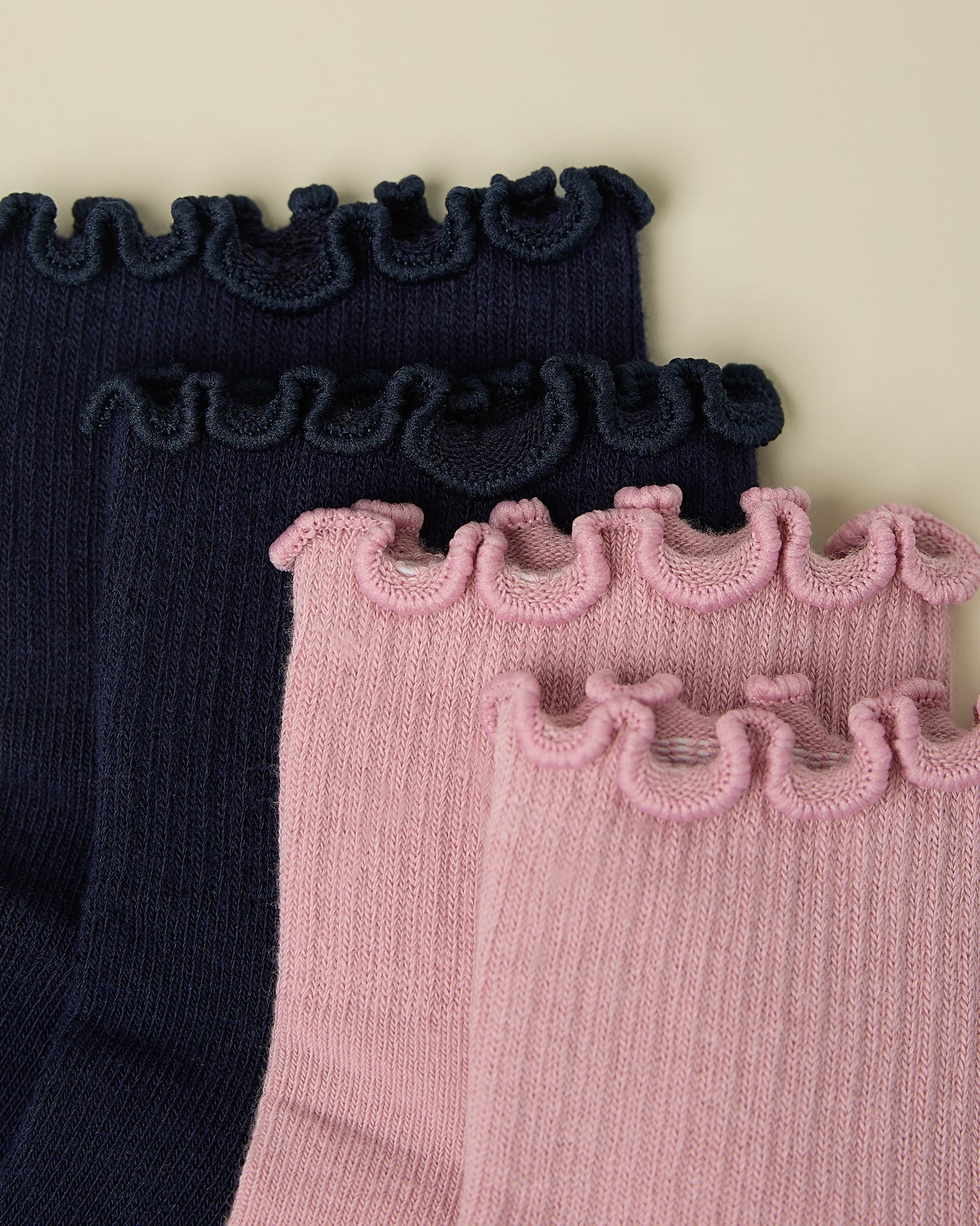 Close up of the top ruffles of two pair of Non-Slip Ruffle Ankle Socks in Navy and Light Mauve on a beige background.