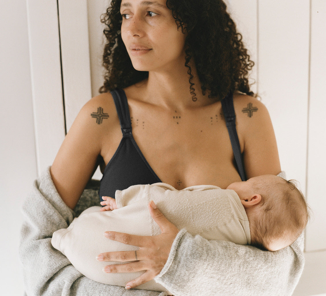 Woman breastfeeding baby in Ribbed Nursing Bra in Black, and Ultra Soft Maternity & Postpartum Cardigan in Grey Heather on a white wall background. Spring Clearance Hero image.