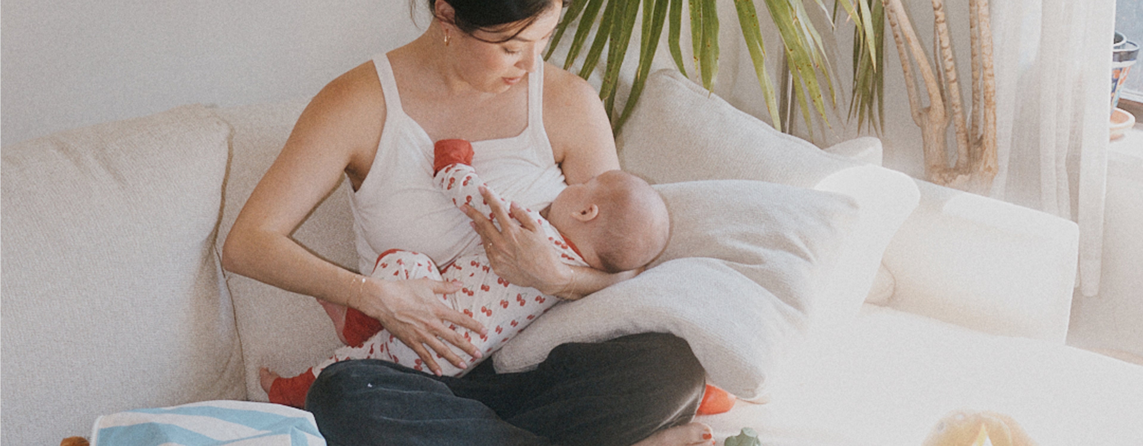 Mother breastfeeding baby on white couch with palm frond and white wall background. Baby is wearing Bamboo Baby Pajama in Ivory Cherries. 