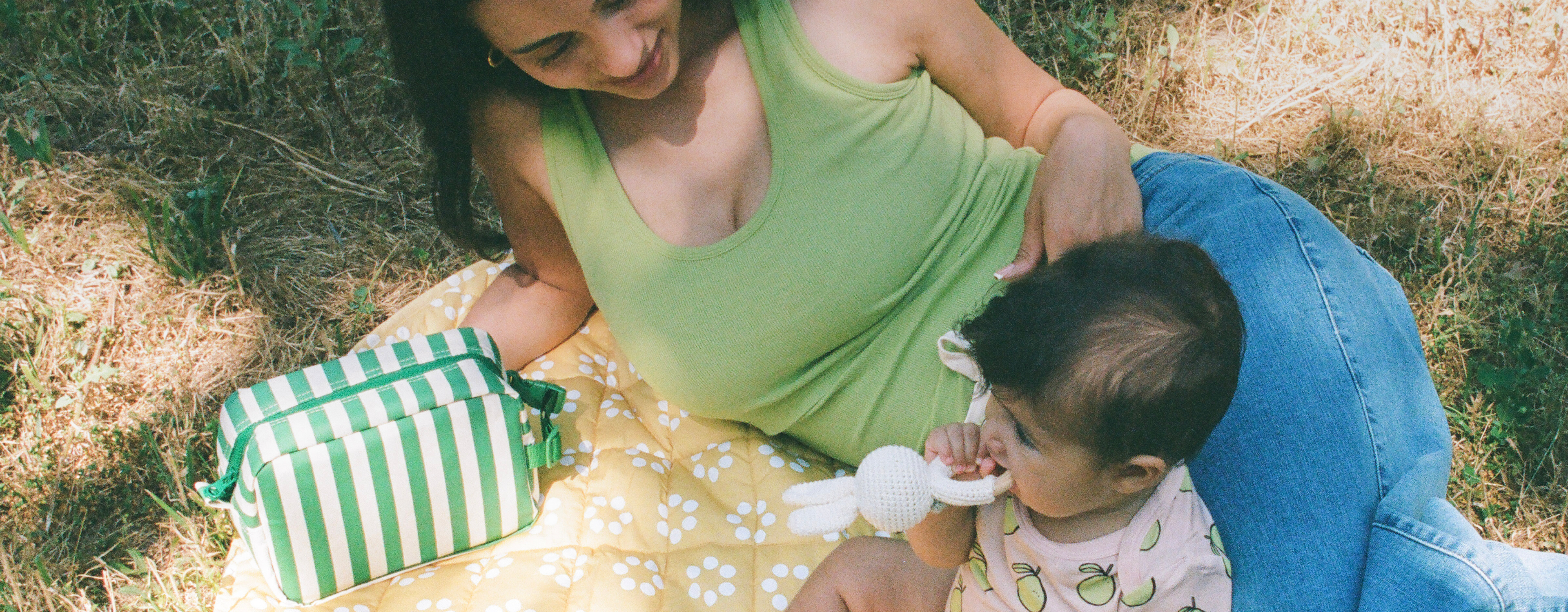 Mom laying on Inside/Outside Play Mat in Sunshine Dots and wearing Ribbed Bamboo Scoop Neck Nursing Tank in Avocado next to baby wearing Bamboo Short Sleeve & Shorts Set in Pink Pomelo on a grassy patch. Next to mom is Mini Cooler Bag in Green Stripe.@model_info:Zoe is wearing a Small.