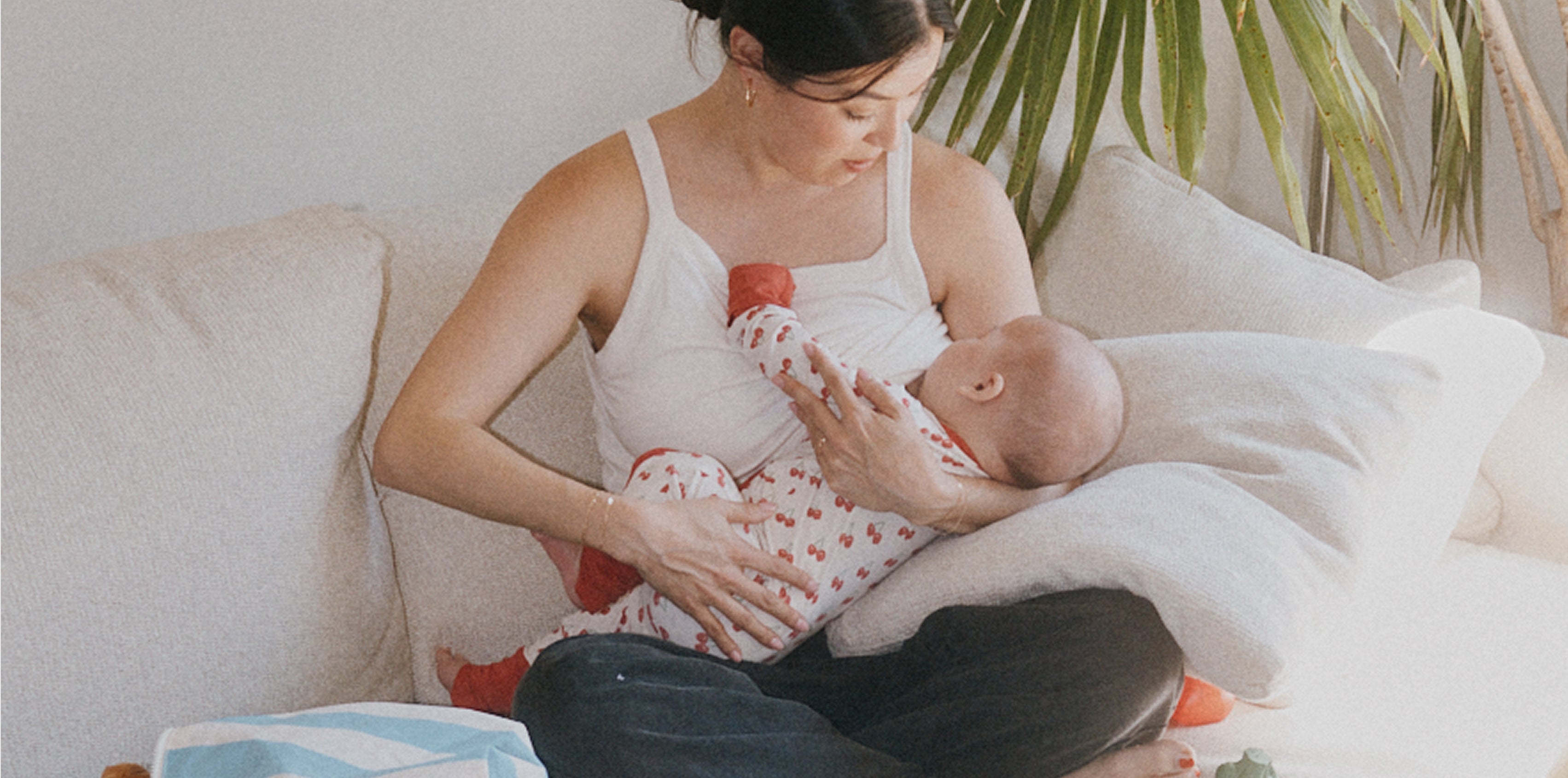 Mother breastfeeding baby on white couch with palm frond and white wall background. Baby is wearing Bamboo Baby Pajama in Ivory Cherries. 