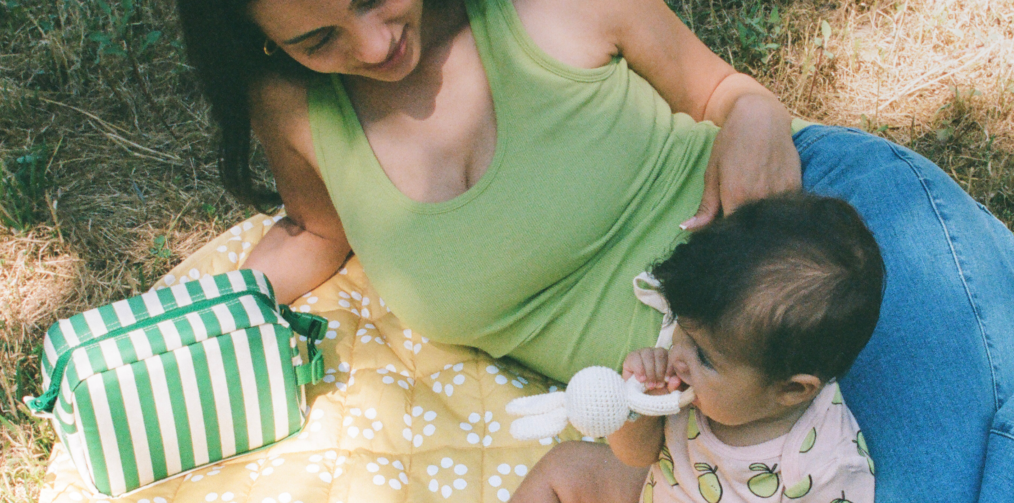 Mom laying on Inside/Outside Play Mat in Sunshine Dots and wearing Ribbed Bamboo Scoop Neck Nursing Tank in Avocado next to baby wearing Bamboo Short Sleeve & Shorts Set in Pink Pomelo on a grassy patch. Next to mom is Mini Cooler Bag in Green Stripe.@model_info:Zoe is wearing a Small.