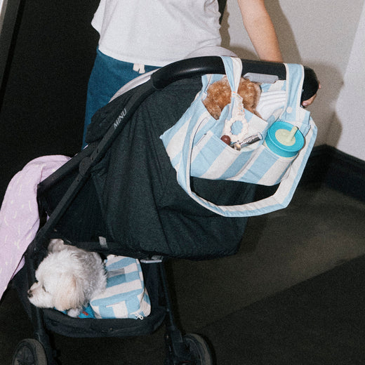 Woman and stroller at elevator, while she looks at baby who is out of view, and covered in Organic Muslin Swaddle in Lilac Squiggles. Stroller holds Stroller Party Caddy Bag in Blue Wide Stripe, full of baby essentials, Mini Cooler Bag in Blue Wide Stripe and a puppy. The woman is wearing Modal Signature Nursing Tank in White. 