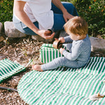 Mommy sitting next to baby on an Inside/Outside Play Mat, part of the On the Go Bundle in Green Stripe, with lush park background.