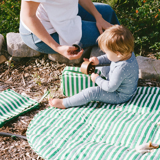 Mommy sitting next to baby on an Inside/Outside Play Mat, part of the On the Go Bundle in Green Stripe, with lush park background.