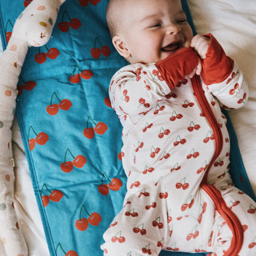 Smiling baby wearing Bamboo Baby Pajama | Ivory Cherries, laying on Quick Change Kit in Blue Cherries. Next to baby is Wet/Dry Bag Set in Stripe, doll and white bed background.@model_info:Johnny is wearing 3-6 months.