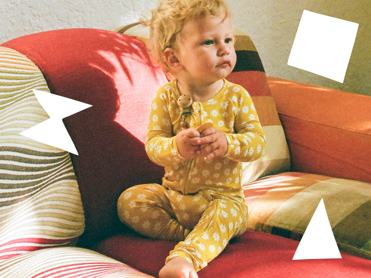 Baby in Bamboo Baby Pajama in Yellow Daisies on a patterned couch with playful white shapes floating around the baby.