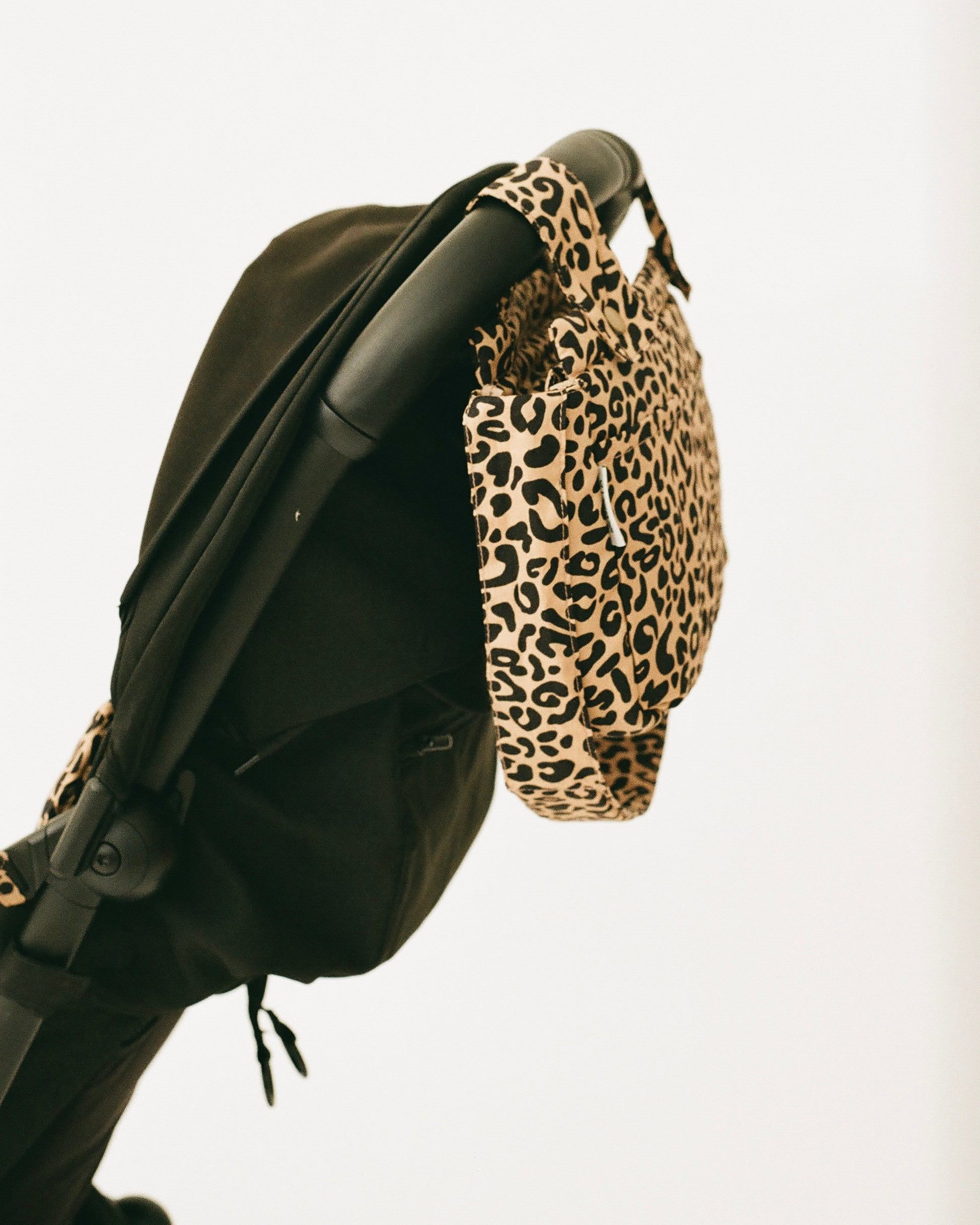 The Stroller Party™ Caddy Bag in Leopard hangs from the handle bar of a black stroller, on a beige background.
