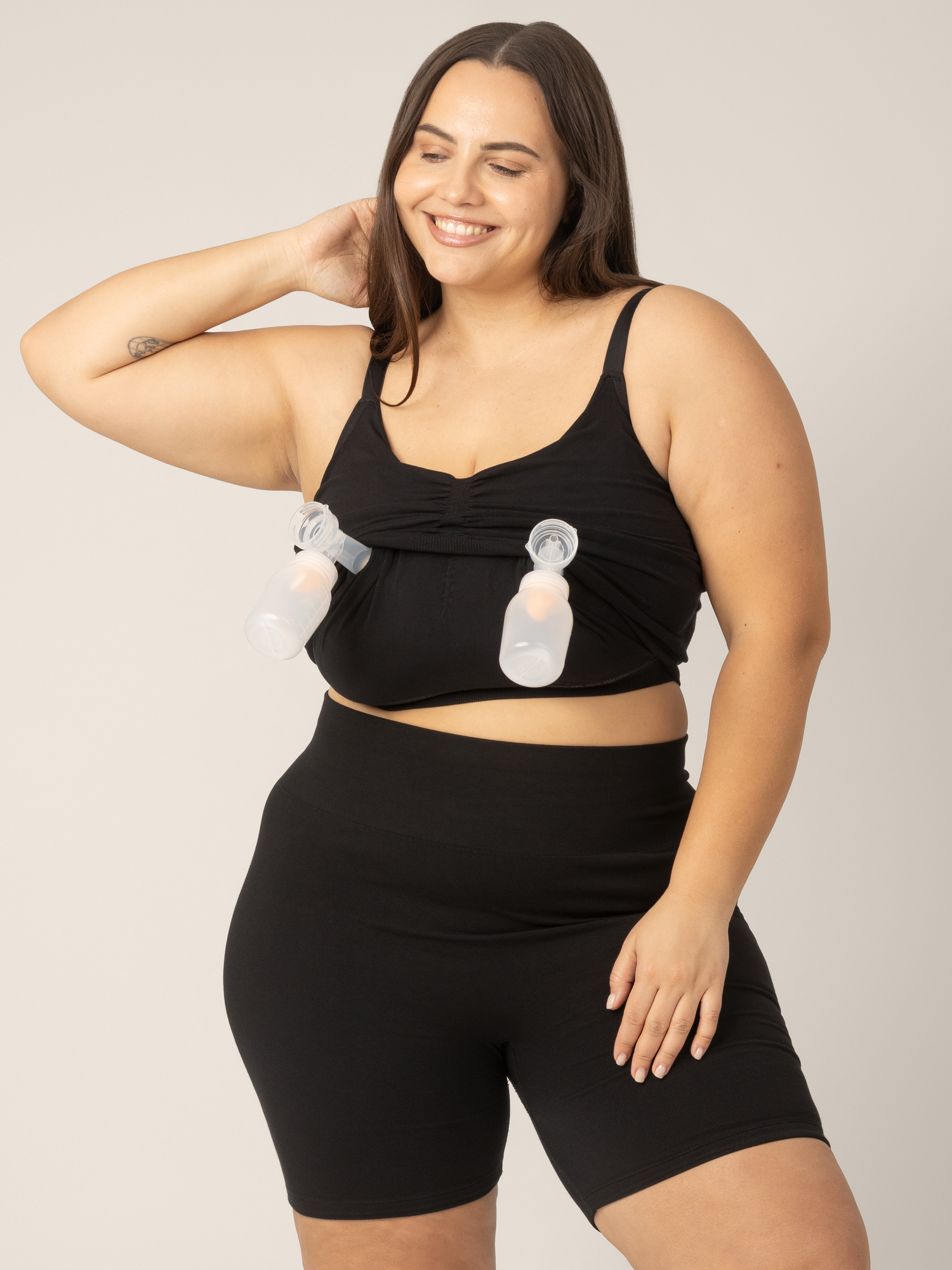 Super busty model wearing the Sublime Hands-Free Pumping & Nursing Sleep and Lounge Bra in Black. Outer layer pulled up to show pumping layer with pump inserted for hands-free pumping access.