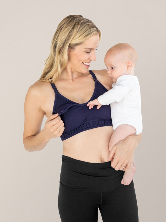Model wearing the Sublime® Hands-Free Pumping & Nursing Sports Bra | Navy holding her baby on her hip and smiling. @model_info:Mia is wearing a Small.
