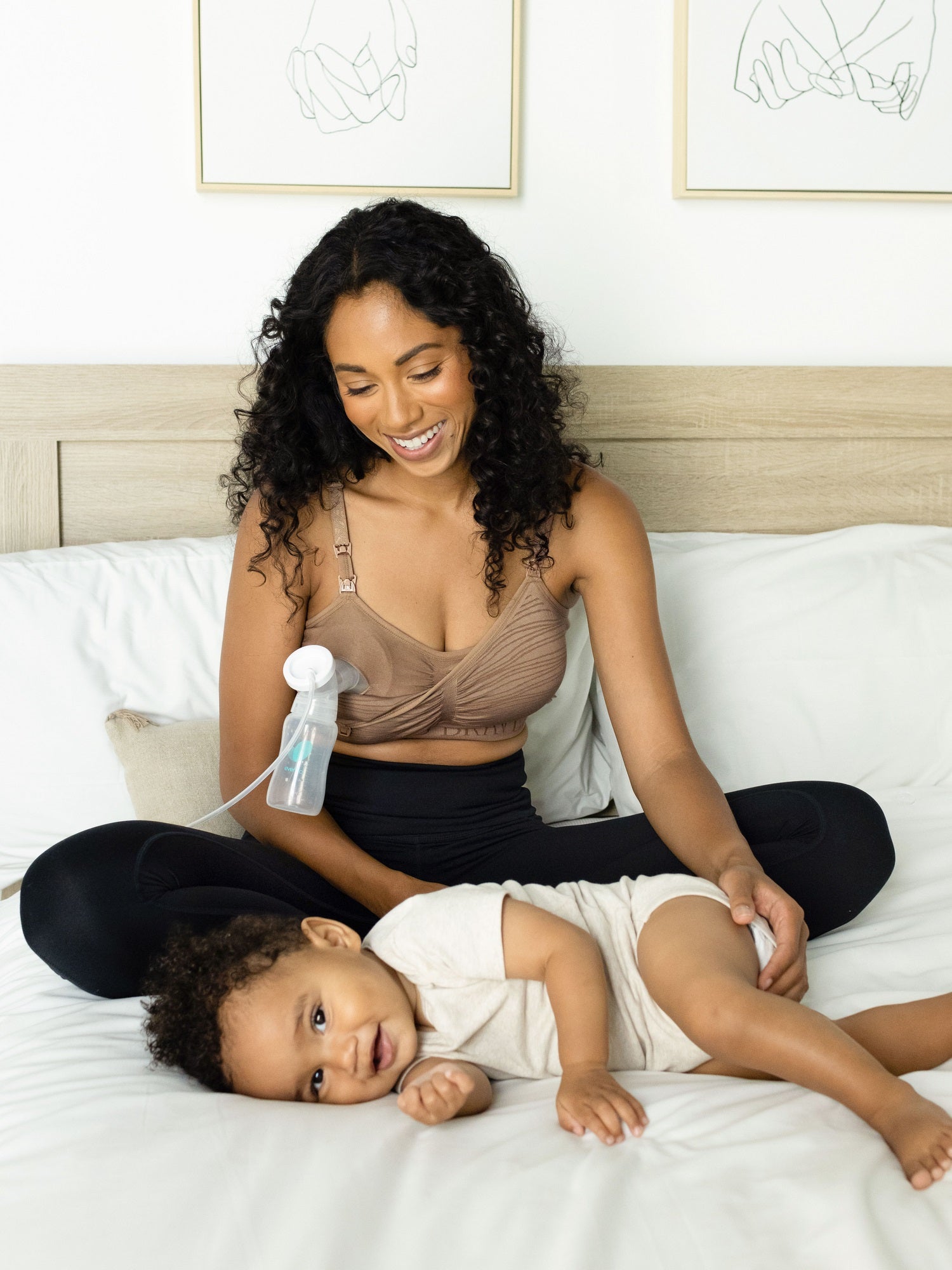 Pumping model sitting on a bed with her baby while wearing the Sublime® Hands-Free Pumping & Nursing Bra in latte@model_info:Anastacia is wearing a Small.