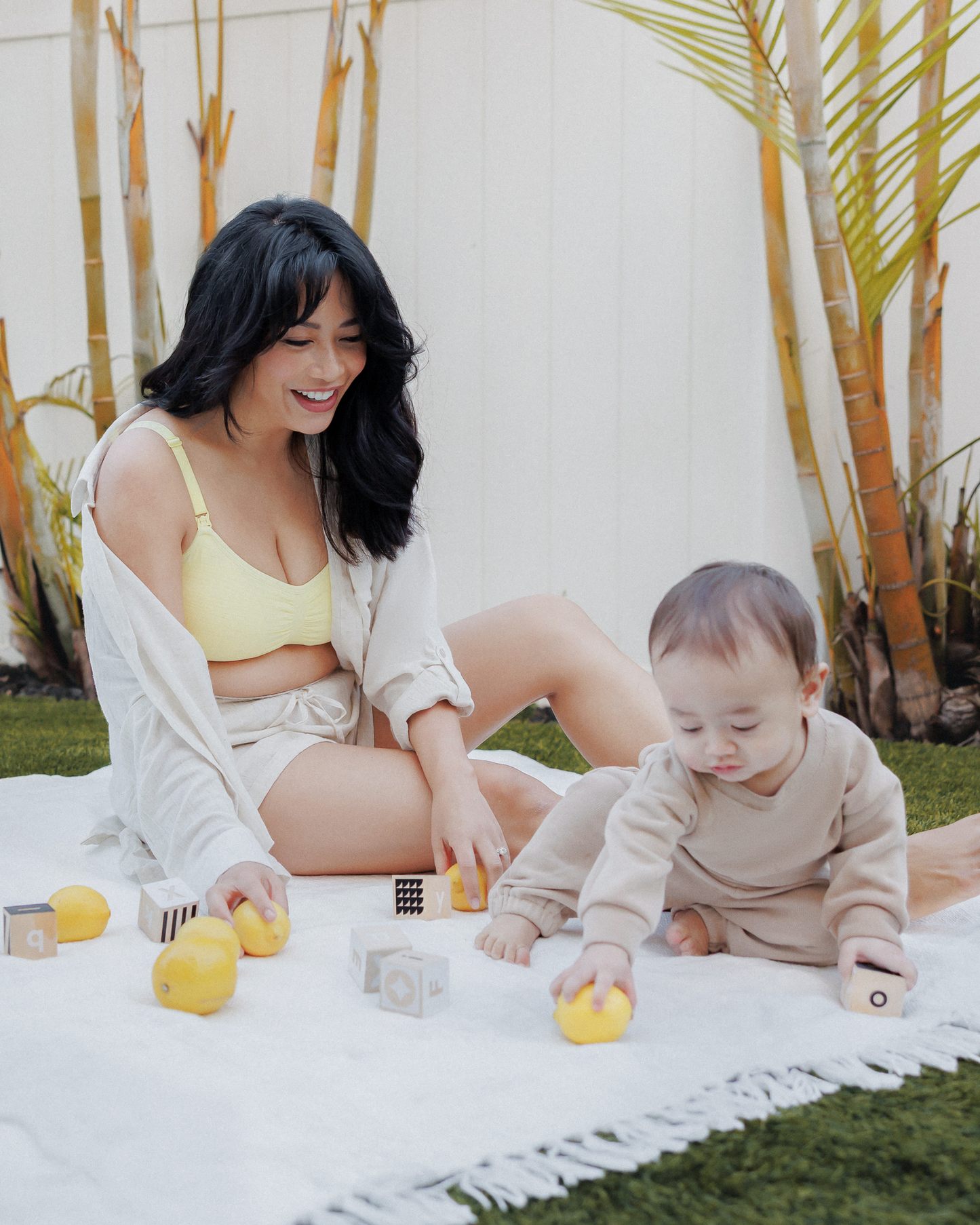 Model outside on blanket on a lawn with baby, wearing the Sublime® Hands-Free Pumping & Nursing Bra Light Lemon, with unbuttoned Reese Linen Oversized Shirt and Reese Linen Shorts @model_info:Nikki is wearing a Medium.