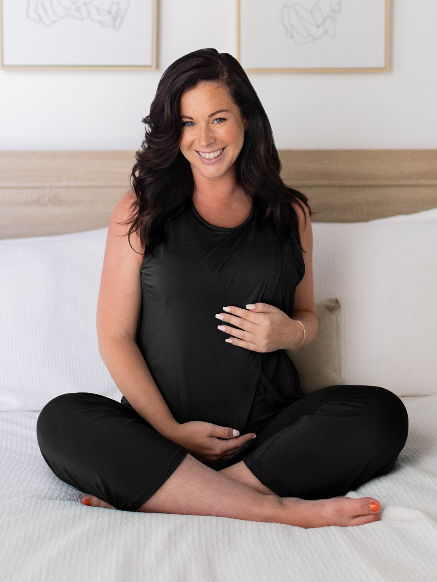 Front view of pregnant model sitting cross-legged on bed wearing the Tulip Hem Nursing Tank & Capri Pajama Set in Black.