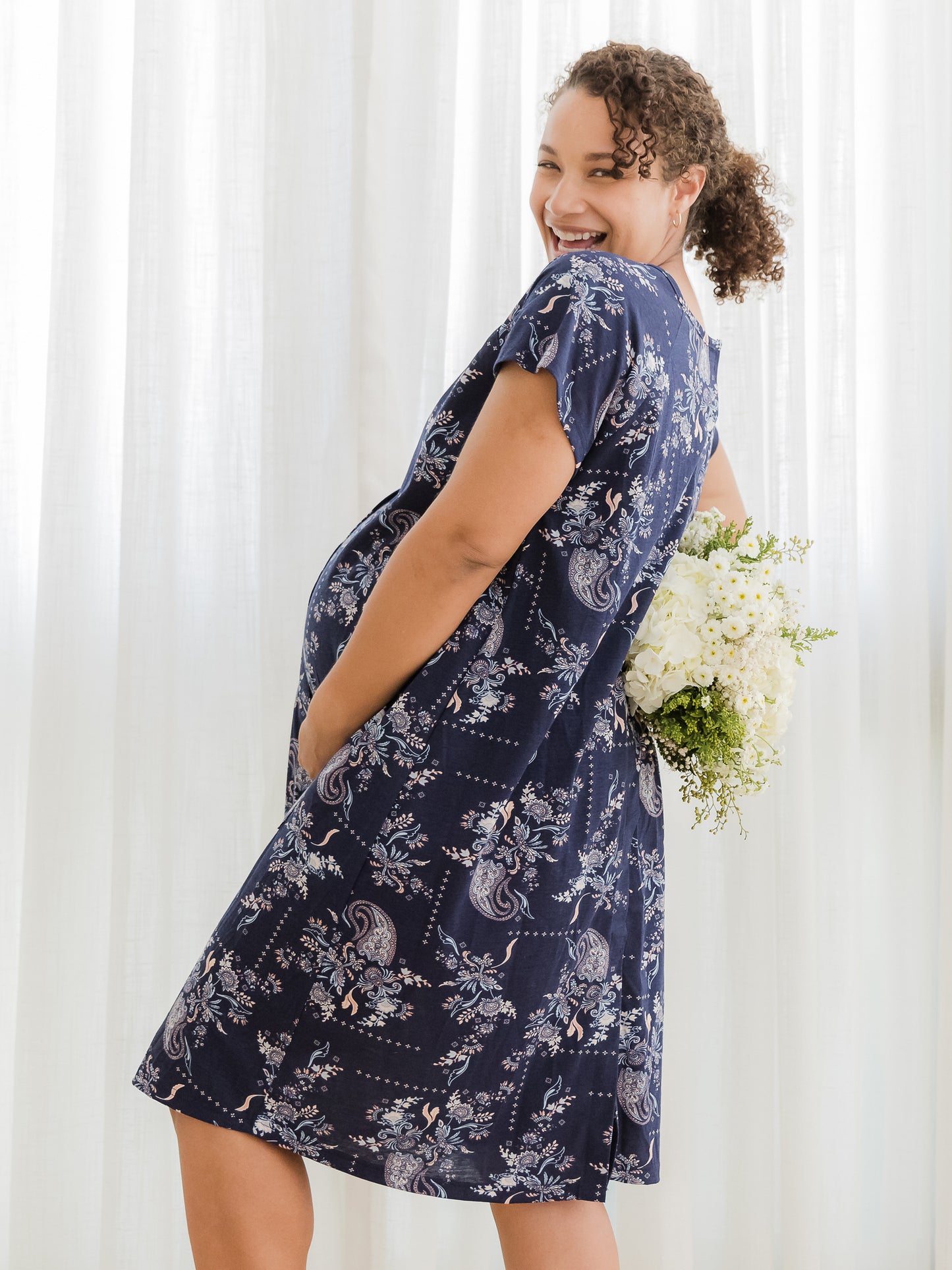 Side view of pregnant model wearing the Universal Labor & Delivery Gown in Navy Paisley, holding flowers in one hand behind her back