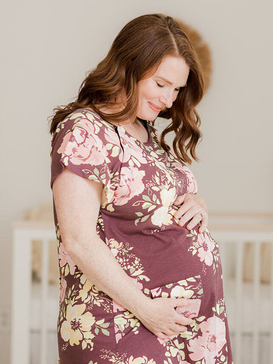 Model wearing the Universal Labor & Delivery Gown in Burgundy Plum Floral with her hands on her pregnant belly standing in front of a crib. @model_info:Amy is wearing a S/M/L.