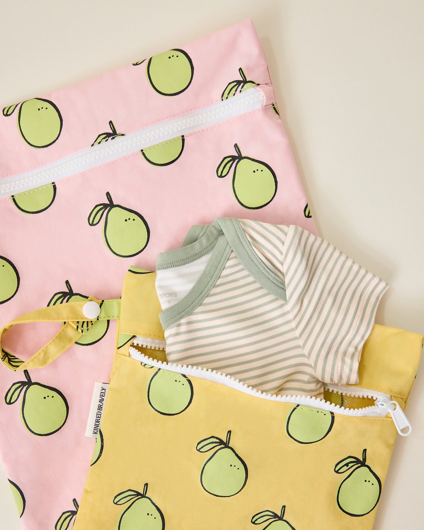 Close up of Wet/Dry Bag Set in Pomelo print unzipped with striped onesie poking out, on beige background