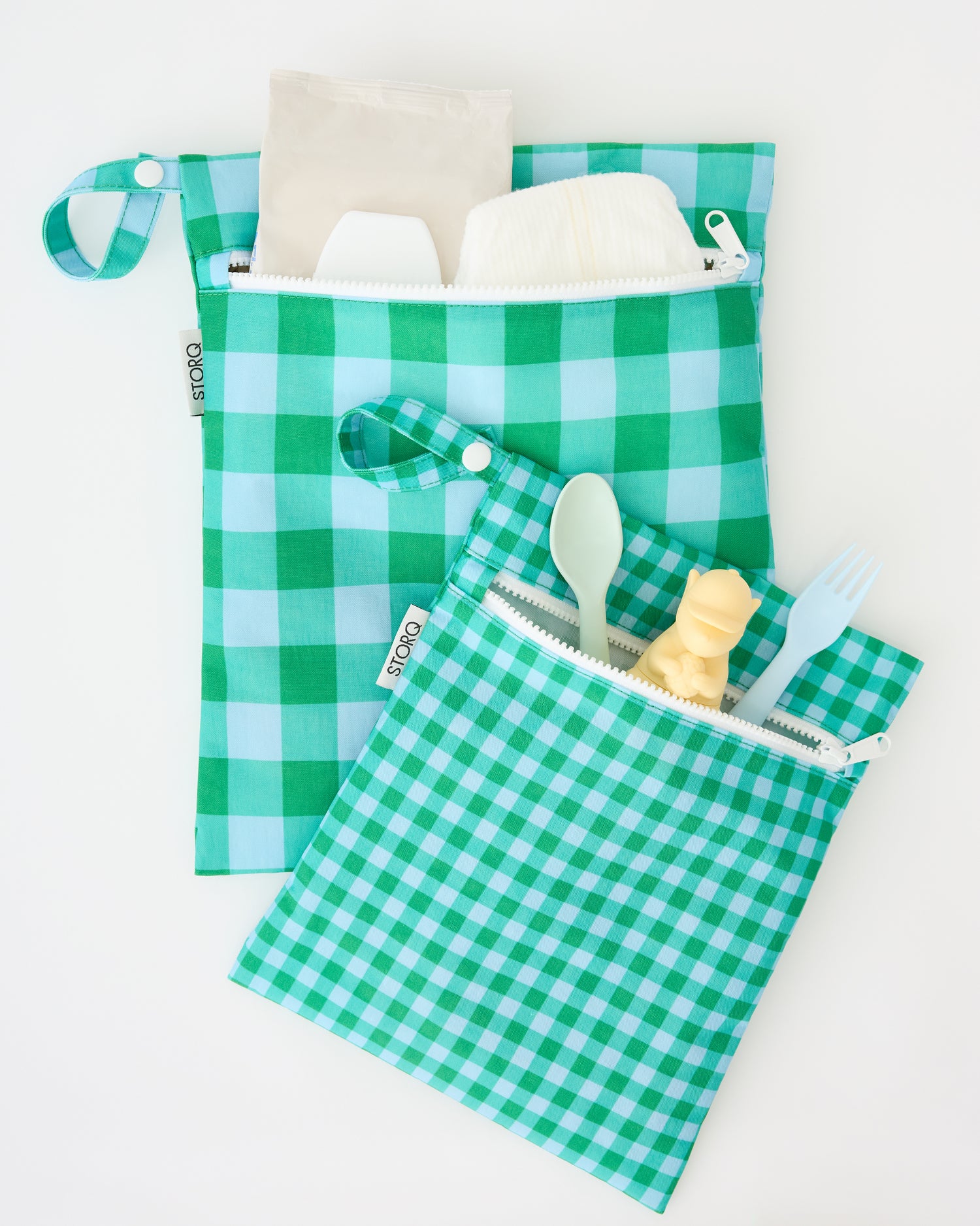 A close up of the openings with diapers, wipes, a chew toy and cutlery peeking out of the Wet/Dry Bag Set in Teal Gingham on a beige background. 