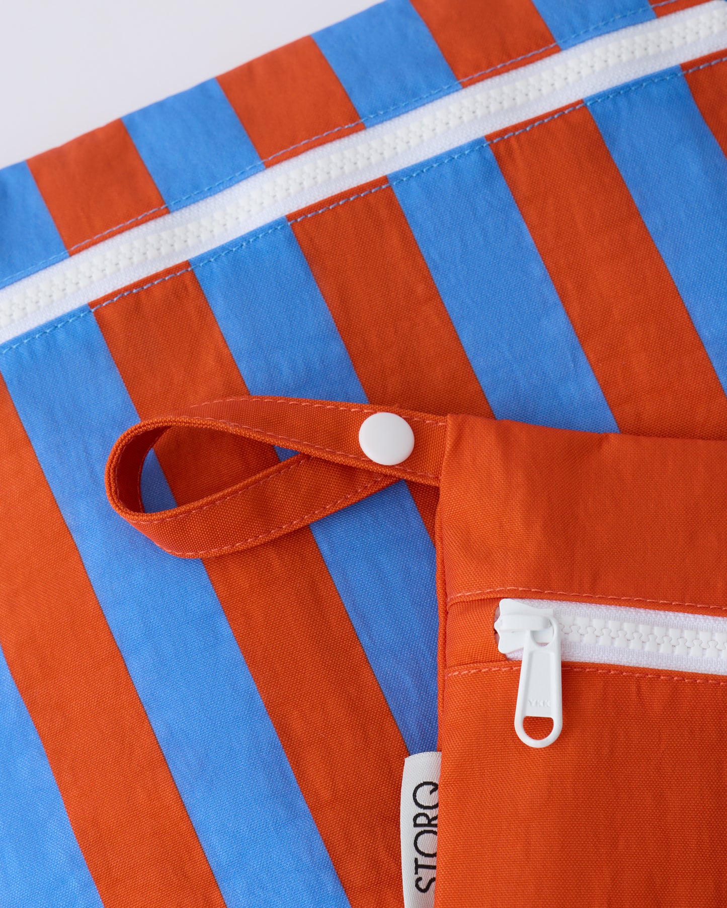 Close up of the zippers and snap loop of the Wet/Dry Bag Set in Blue & Rust Stripe on a beige background.