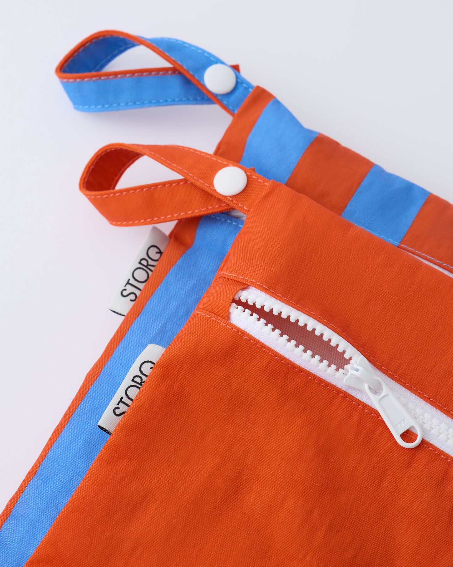 Close up of the zippers and snap loop of the Wet/Dry Bag Set in Blue & Rust Stripe on a beige background.