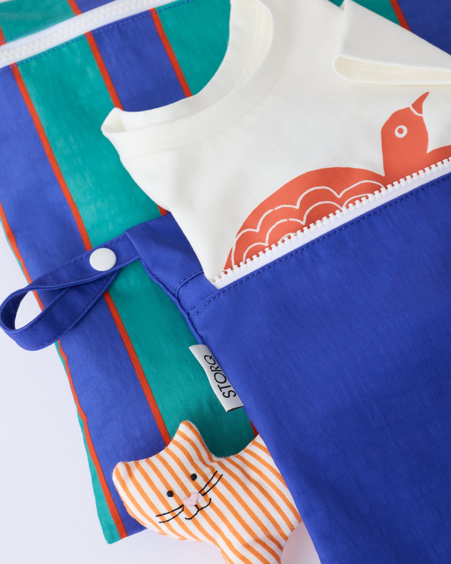 Close up of Wet/Dry Bag Set in Navy & Green Stripe with a shirt sticking out of the smaller bag, and a toy underneath, on a beige background.