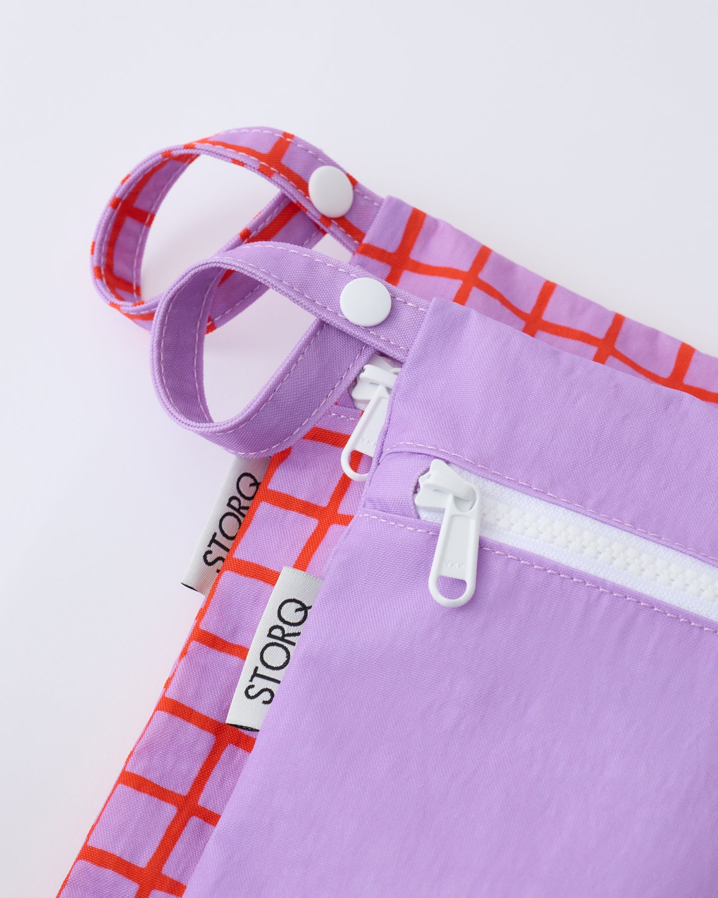 Close up of the zippers and snap loop of the Wet/Dry Bag Set in Orchid Grid on a beige background.