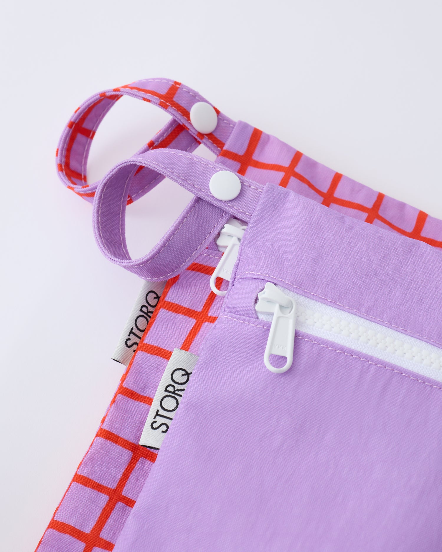 Close up of the zippers and snap loop of the Wet/Dry Bag Set in Orchid Grid on a beige background.