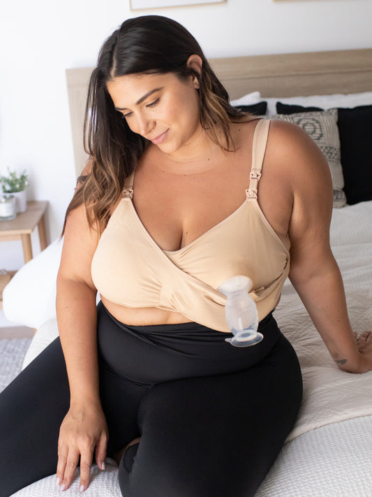 Busty model wearing the Sublime® Hands-Free Pumping & Nursing Bra - Super Busty in Beige sitting on bed with one pump flange in@model_info:Gia is wearing a 1X Super Busty.