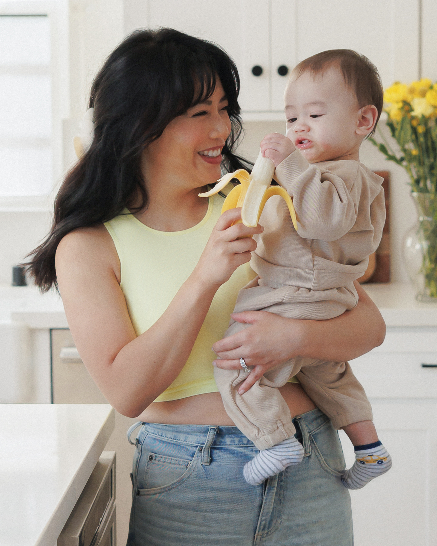 Model standing in kitchen holding baby and wearing the Sublime® Bamboo Maternity & Nursing Longline Bra Top in Light Lemon, paired with jeans @model_info:Nikki is wearing a Mediium.