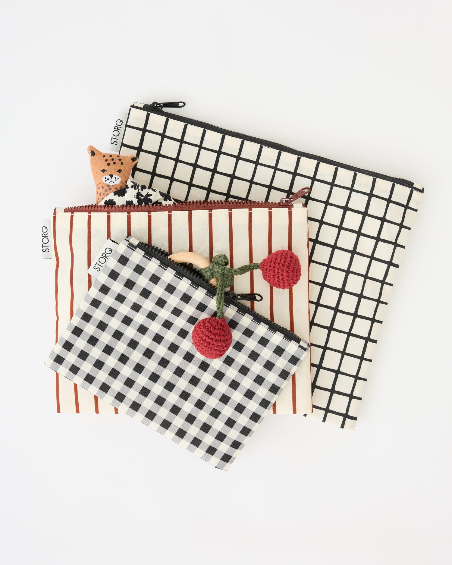 Zipper Pouch Three-Piece Set in Black Grid with a woven cherry chew toy and a leopard lovey doll sticking out of the pouches, on a beige background.