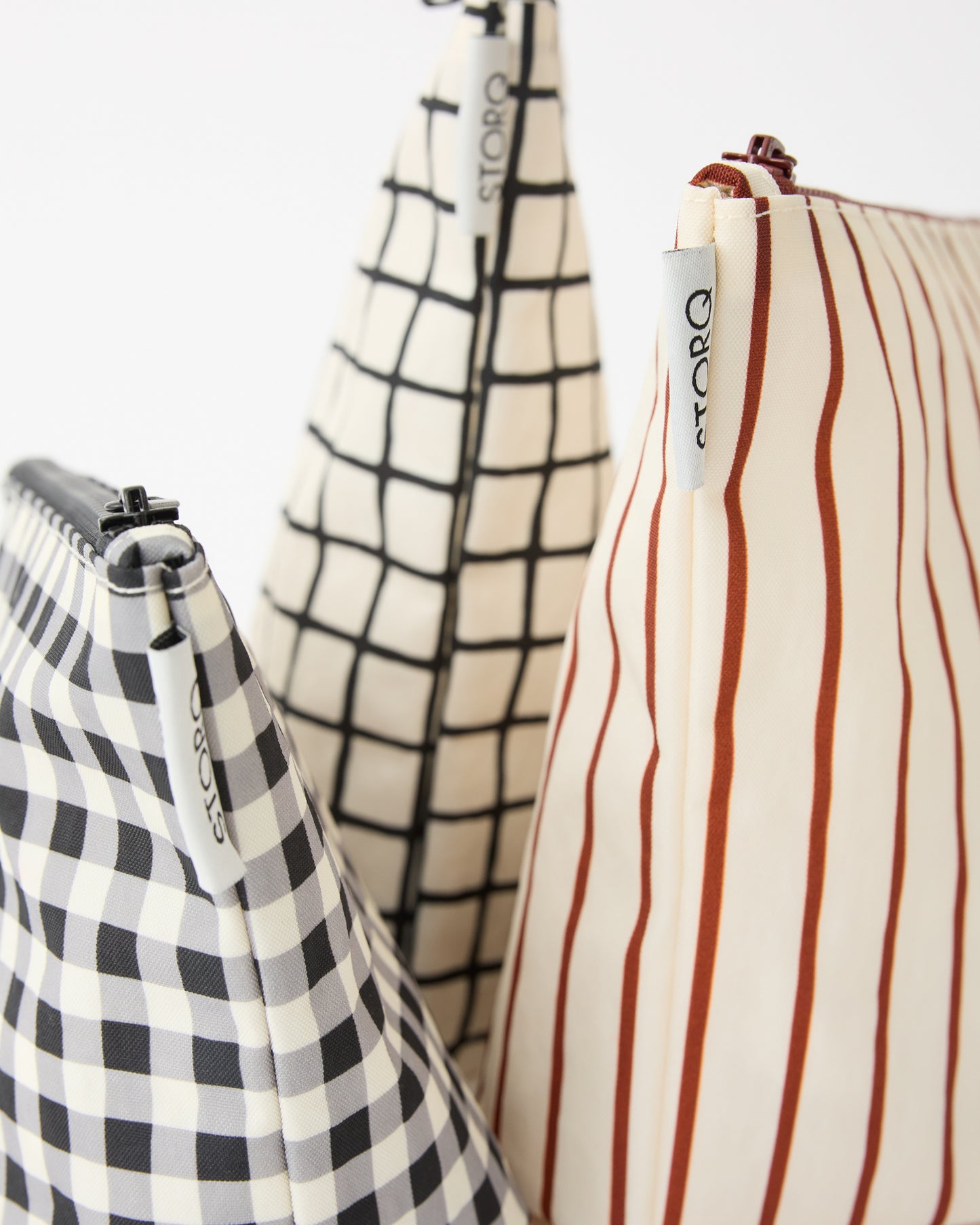 Close up of the zippers, tags and various patterns of the Zipper Pouch Three-Piece Set in Black Grid on a beige background.