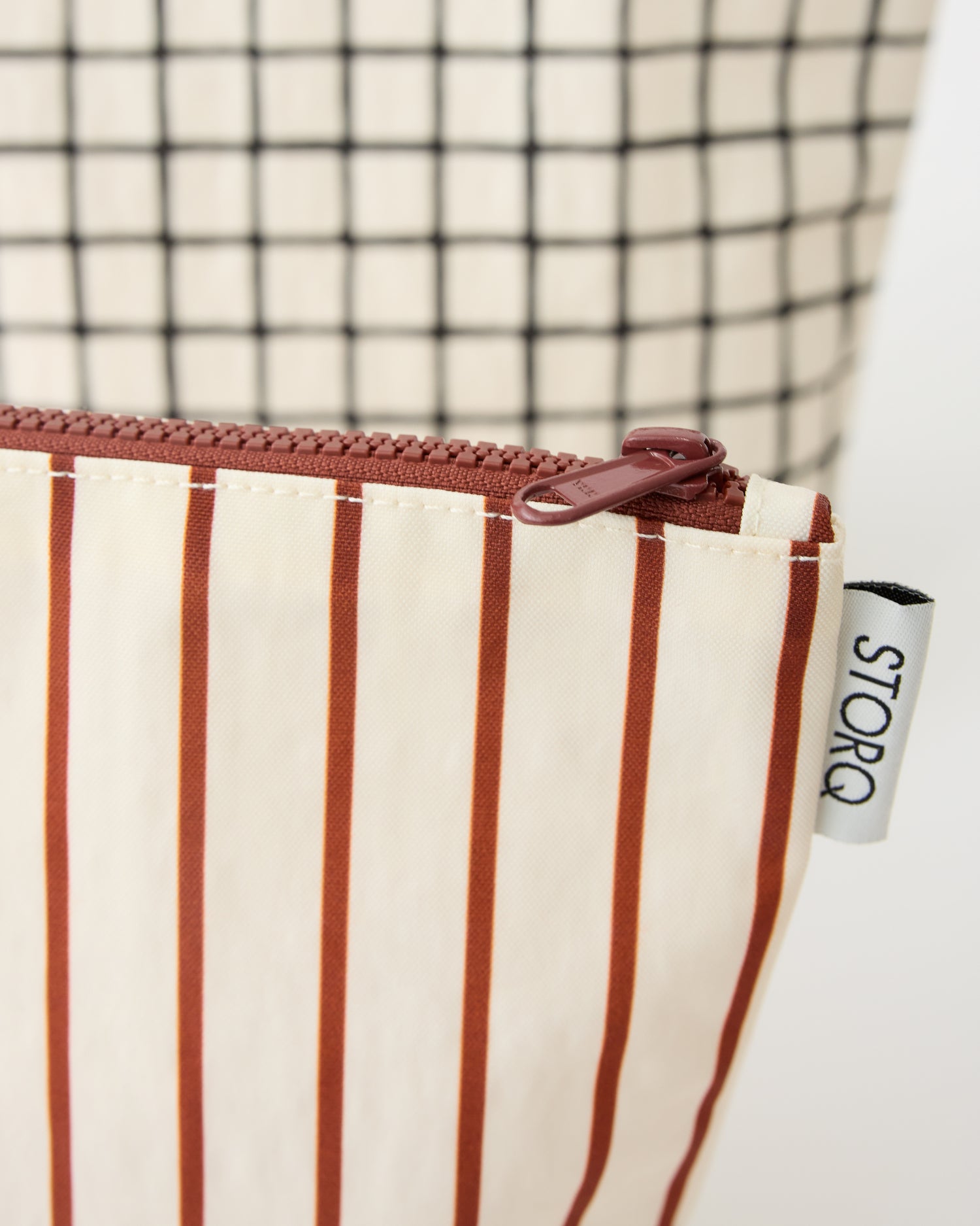 A close up at the pattern, tag and zipper of the Zipper Pouch Three-Piece Set in Black Grid on a beige background.