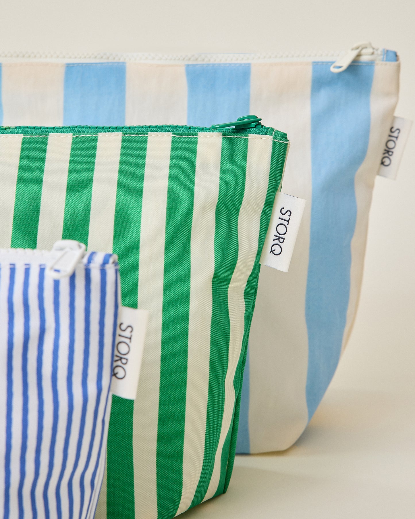 Close up of the wide blue stripe, wide green stripe and thin blue stripe pouches of the Zipper Pouch Three-Piece Set in Stripe print on a beige background