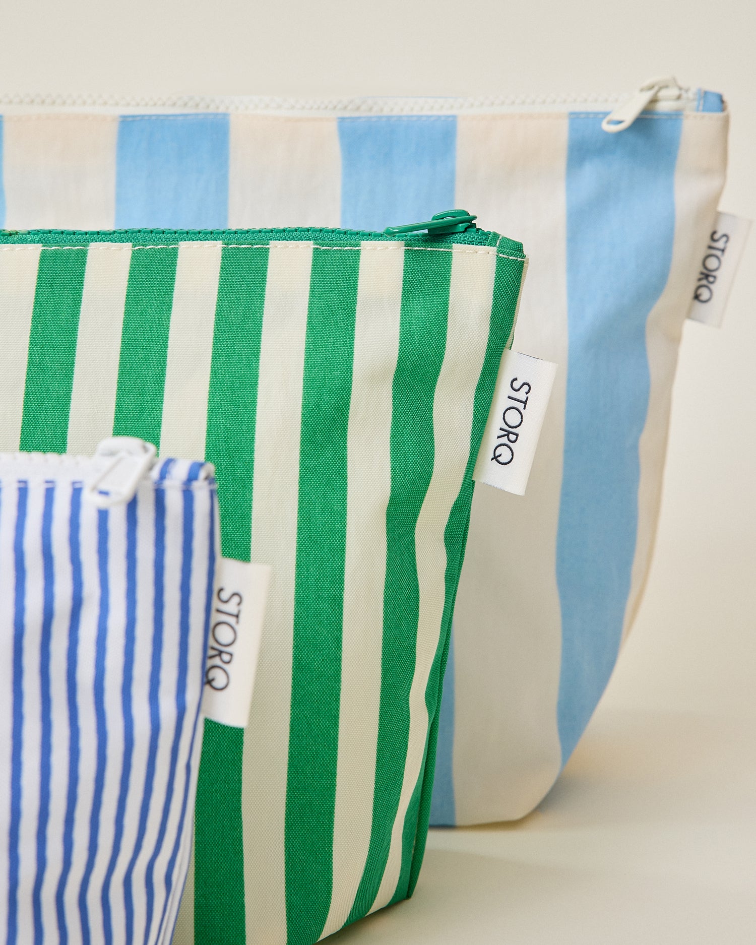 Close up of the wide blue stripe, wide green stripe and thin blue stripe pouches of the Zipper Pouch Three-Piece Set in Stripe print on a beige background