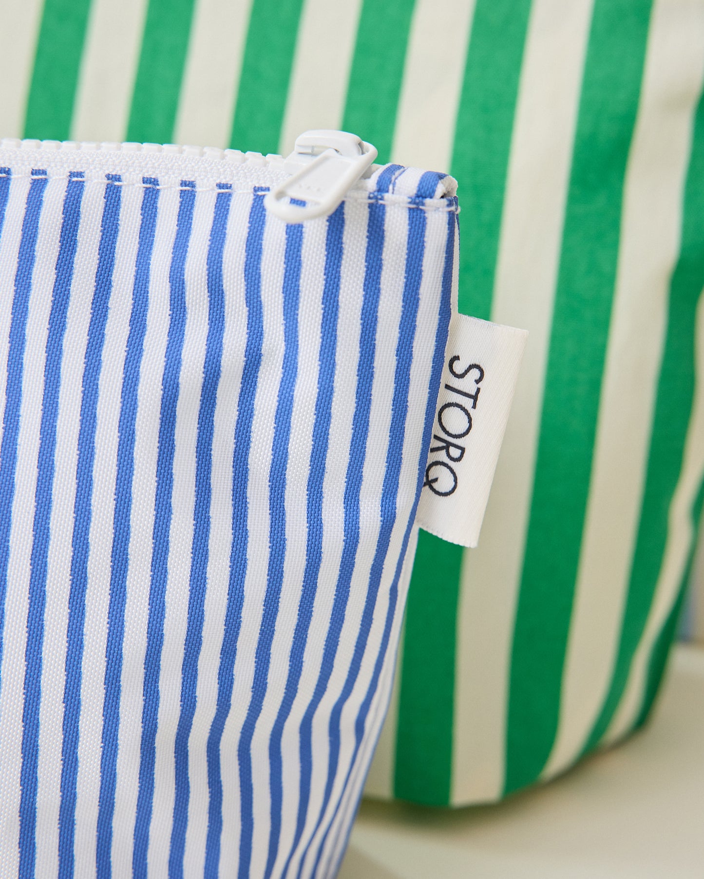 Close up of the thin blue stripe and wide green stripe pouches of the Zipper Pouch Three-Piece Set in Stripe print on a beige background