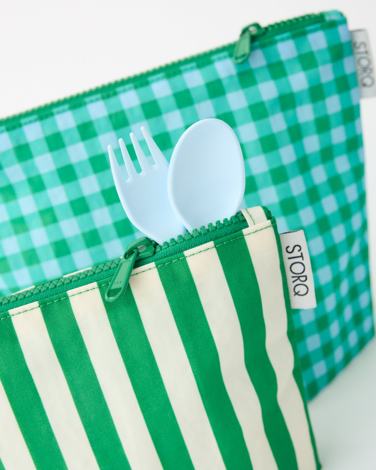 Close up on the zippers, showing a fork and spoon sticking out of the Zipper Pouch Three-Piece Set in Teal Gingham on a beige background.