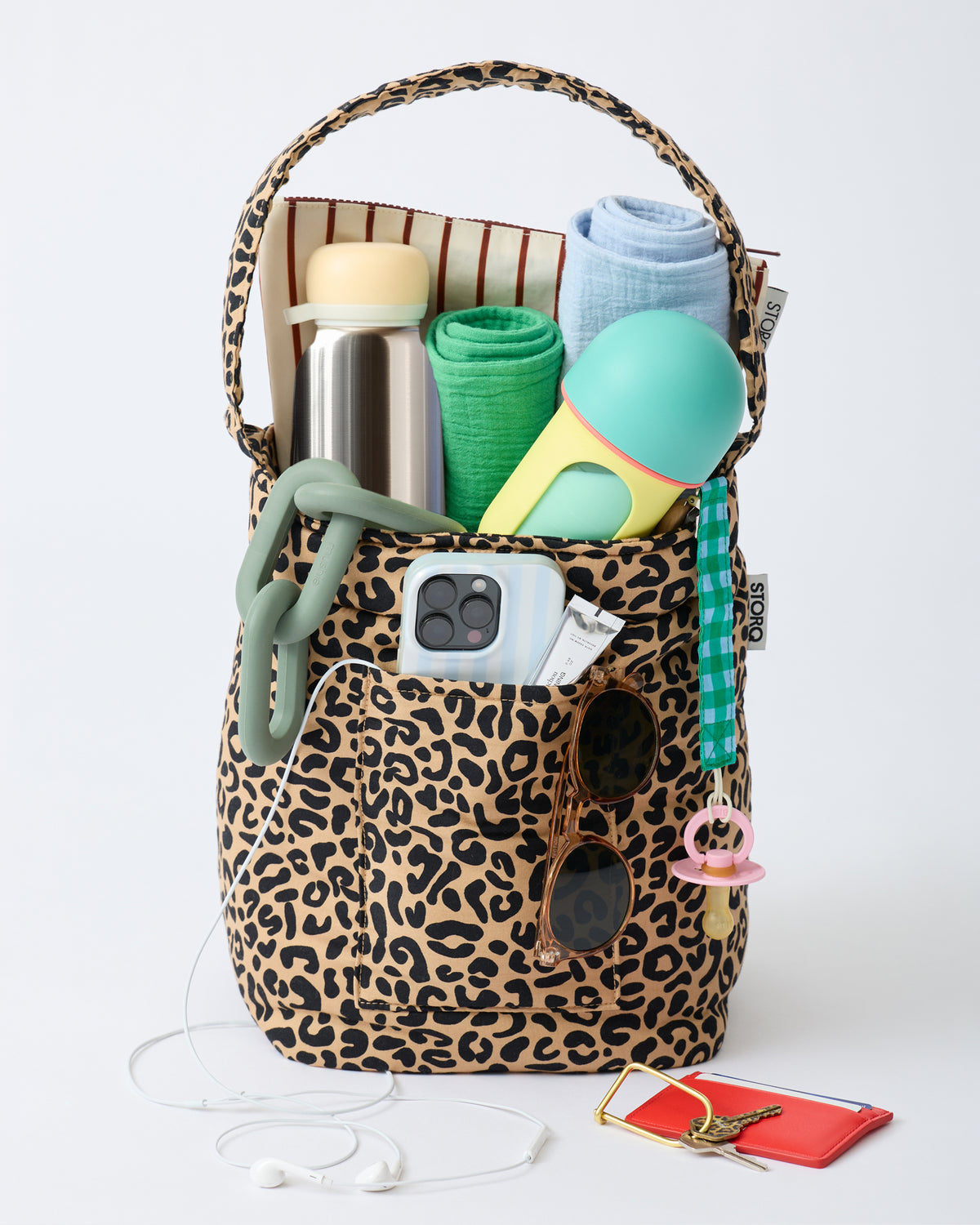 Stroller Party™ Bucket Bag in Leopard is sitting upright, filled with Muslin Cloths, Wet/Dry bag, chewing chain toy, water bottle, milk bottle, phone, shades, lip balm, Pacifier Clip, wallet and keys, on an off white background.