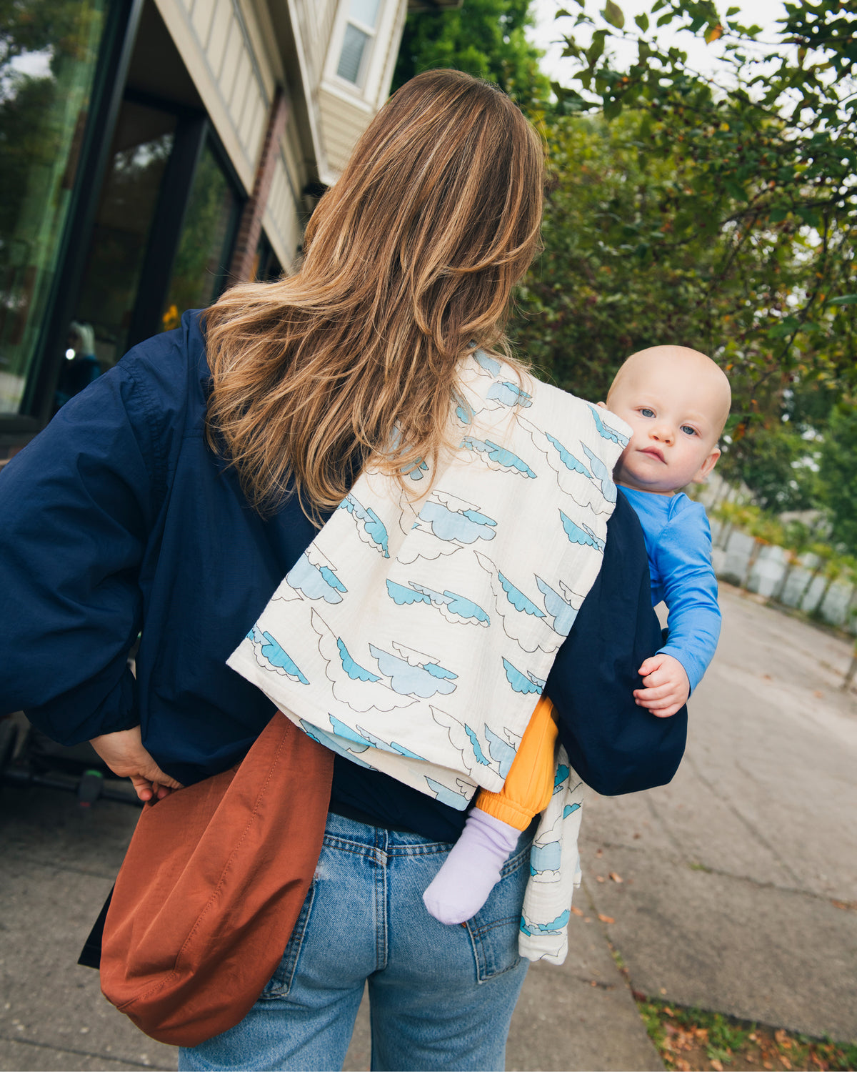 A woman outdoors carrying a baby on her waist and a Organic Muslin Burp Cloth in Ivory Clouds pattern over her shoulder. The woman is wearing a navy blue jacket and jeans, with Quick Change Half Moon Bag in Cinnamon.
