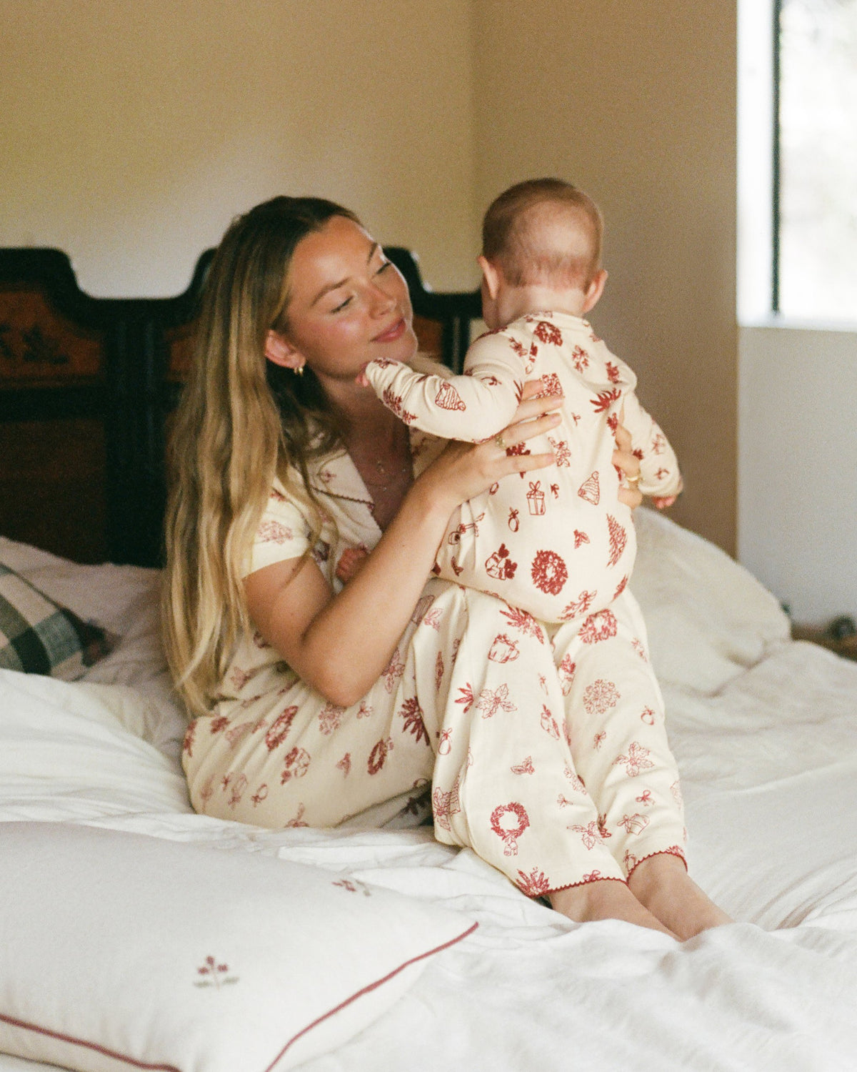 A woman and baby wearing matching holiday-themed pajamas sitting on a bed. Mother is wearing Cotton Bamboo Pajama Set in Cozy Day and baby is wearing Bamboo Baby Pajama in Cozy Day. In a sun lit bedroom.