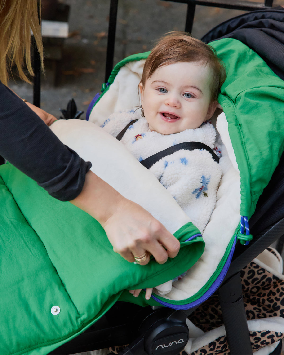 A black stroller has Cozy Stroller Footmuff in Green secured into it with baby sitting inside, while peeking out and smiling while the woman is unzipping the front panel. The baby wears a fuzzy white and blue outfit.