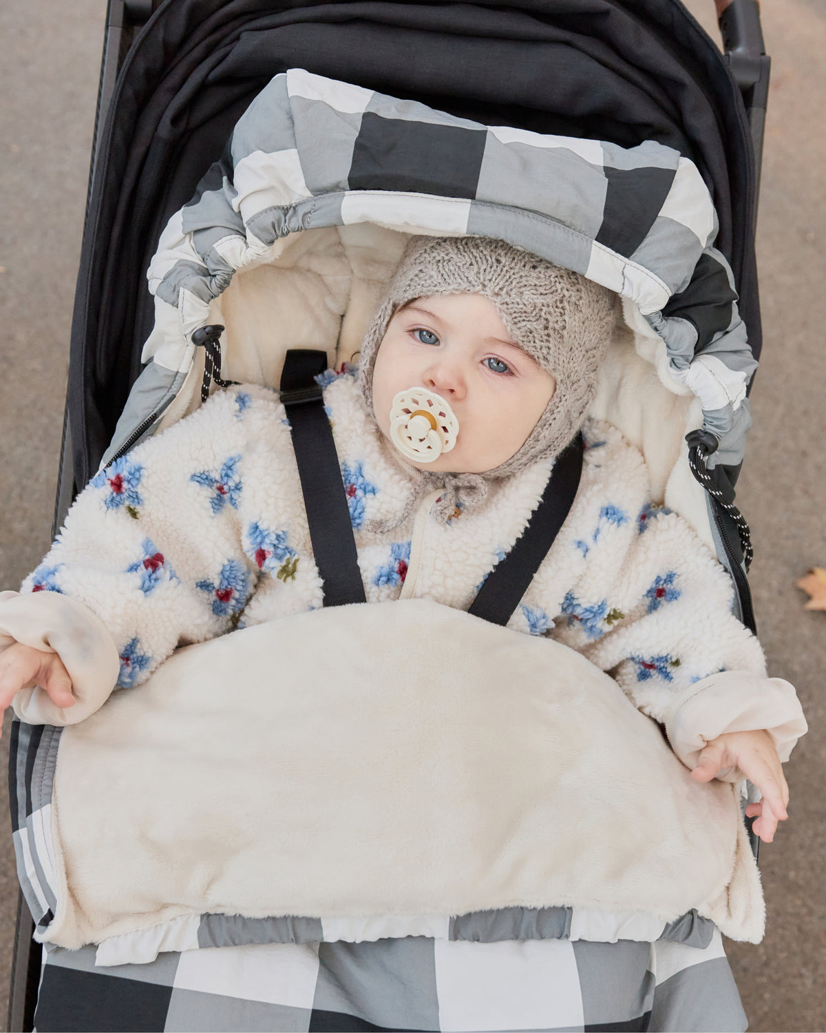A baby wearing a cream sherpa jacket with blue and red floral embroidery, a knit beanie, and a pacifier, sitting in a stroller within a Cozy Stroller Footmuff in Buffalo Plaid, in a city park background.