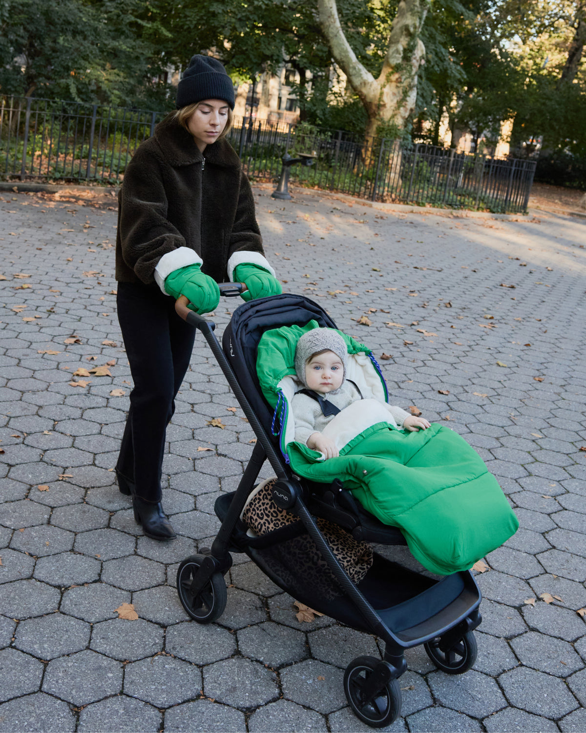 A woman pushes a black stroller while wearing Cozy Stroller Mitts in Green, on a tiled outdoor park path while a child sits inside. The stroller has Cozy Stroller Footmuff in Green secured into it with baby sitting inside, and the child wears a light knit hat and grey outfit.