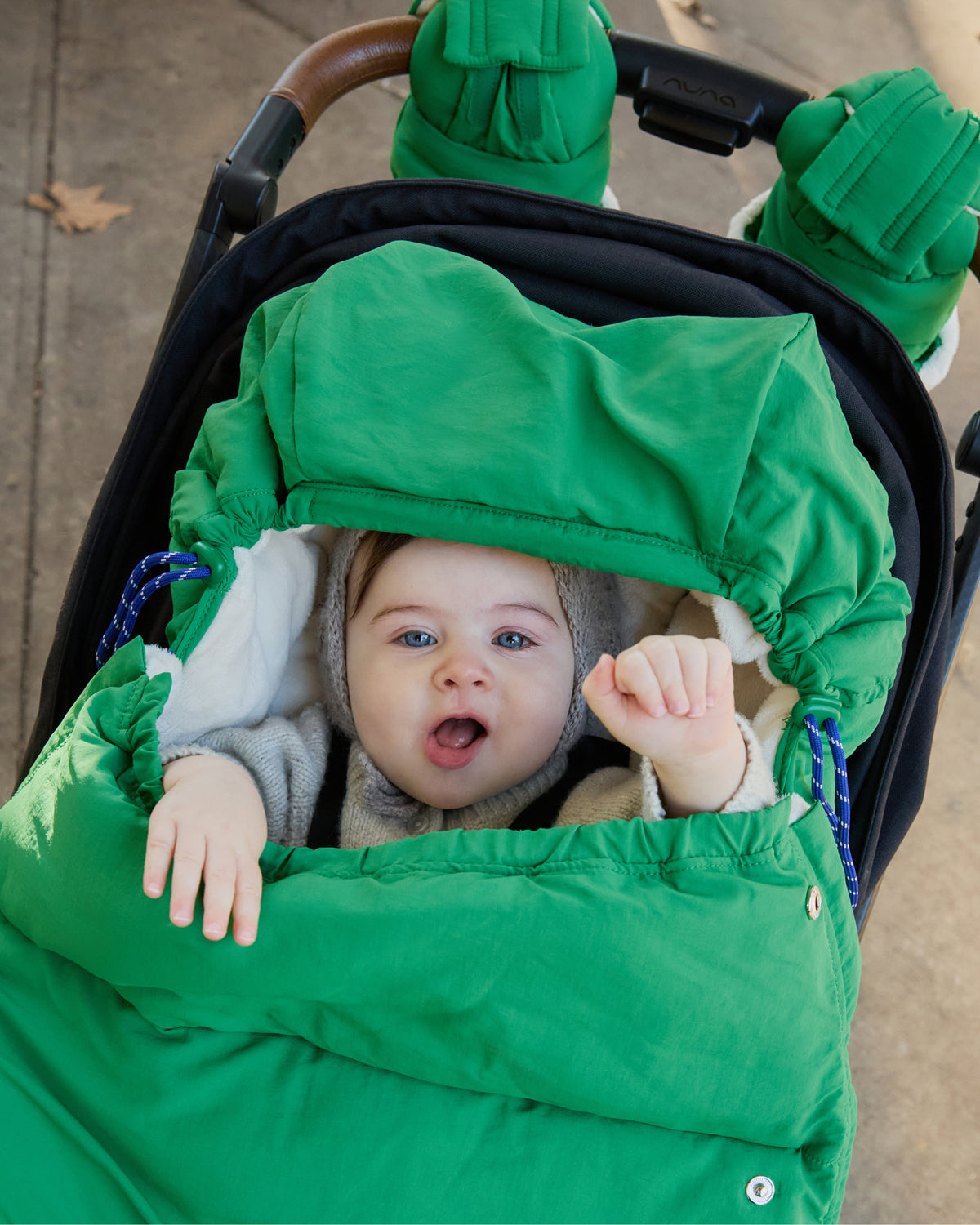 Cozy Stroller Mitts in Green are attached to a black and brown stroller handlebar. The stroller has Cozy Stroller Footmuff in Green secured into it with baby sitting inside, while peeking out and waving. The baby wears a light knit hat and grey outfit.